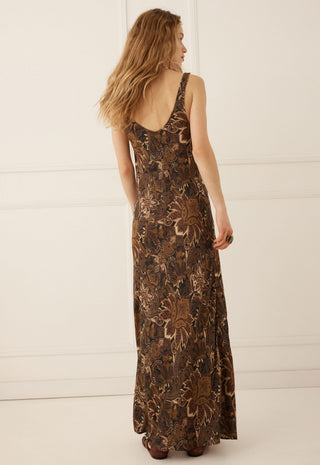 a-woman-wearing-a-sleeveless-floor-length-brown-maxi-dress-with-a-floral-pattern-shown-from-the-back-against-a-white-paneled-wall